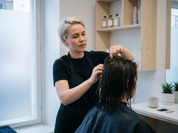 Kampaaja leikkaa asiakkaan hiuksia valoisassa ja minimalistisessa kampaamossa talvipäivän pehmeässä luonnonvalossa