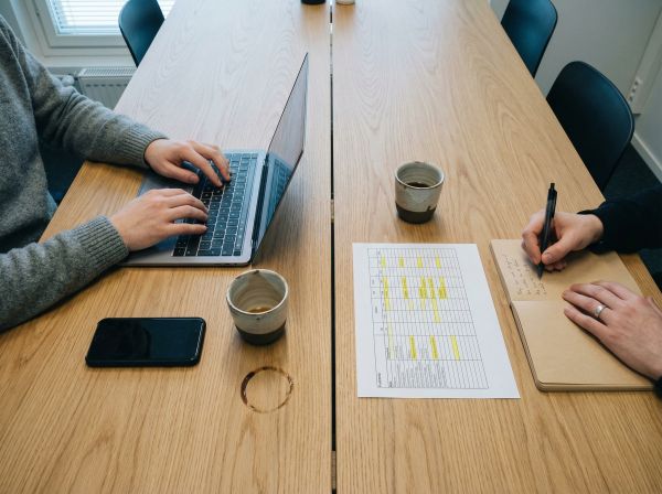 Lähikuva palaverin yksityiskohdista: kädet kannettavalla ja muistikirjalla, kahvikupit ja tuloste vaalealla tammipöydällä