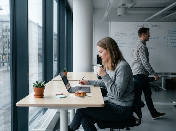 Nainen työskentelee kannettavalla tietokoneella koivuisessa avotoimistossa kahvikuppi kädessään, taustalla liikkuva kollega ja luonnonvalo ikkunasta